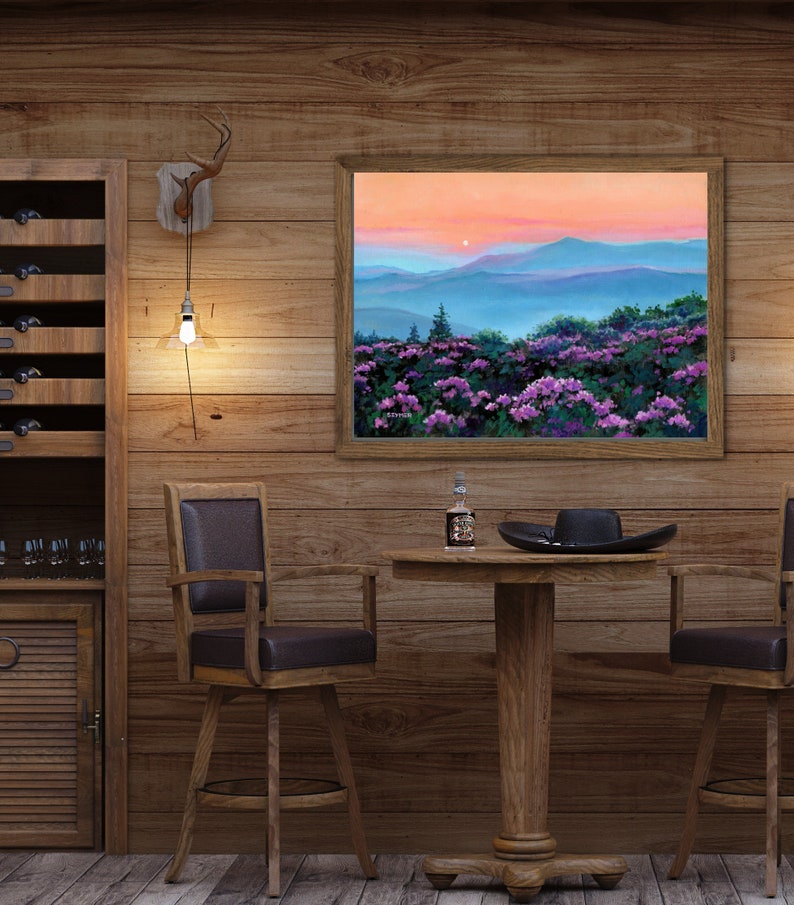 May include: A framed landscape painting depicting a sunset over blue mountain ridges and a foreground of vibrant purple rhododendron flowers. The artwork is displayed on a wood-paneled wall above a round wooden table with a bottle and a black hat.
