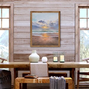 May include: A framed rectangular painting depicting a sunset over a beach with soft clouds, calm ocean waves, and wet sand. The artwork features a warm color palette of gold, orange, and blue tones, encased in a simple light brown wooden frame.