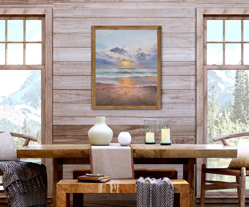 May include: A framed rectangular painting depicting a sunset over a beach with soft clouds, calm ocean waves, and wet sand. The artwork features a warm color palette of gold, orange, and blue tones, encased in a simple light brown wooden frame.
