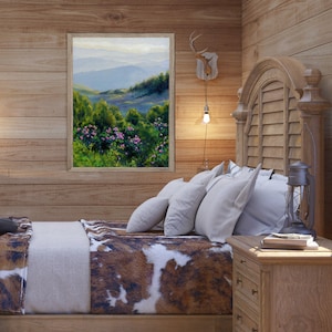 May include: A framed landscape painting depicting a mountain range with green trees and pink flowers in the foreground, displayed on a wood-paneled wall above a bed with a brown and white cowhide-patterned blanket and white pillows.