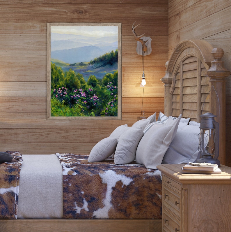 May include: A framed landscape painting depicting a mountain range with green trees and pink flowers in the foreground, displayed on a wood-paneled wall above a bed with a brown and white cowhide-patterned blanket and white pillows.