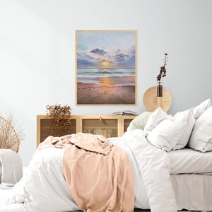 May include: A framed rectangular wall art print depicting a beach scene at sunset with soft blue and purple clouds, gentle ocean waves, and a sandy shore. The artwork is displayed above a wooden cabinet with a light-colored bed in the foreground.