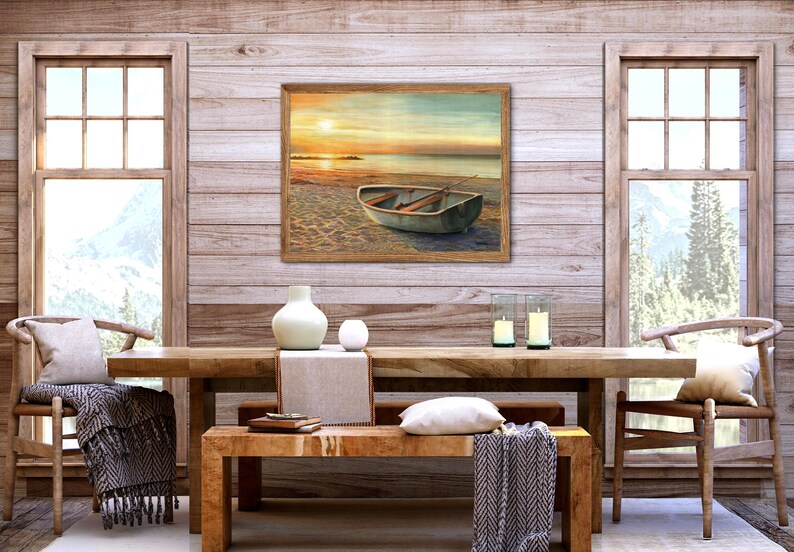 Op de afbeelding: Een rechthoekige ingelijste kunstdruk met een kleine roeiboot op een zandstrand bij zonsondergang, met zacht gouden licht dat reflecteert op het water en de horizon, in een kamer met een houten tafel, stoelen en kaarsen.