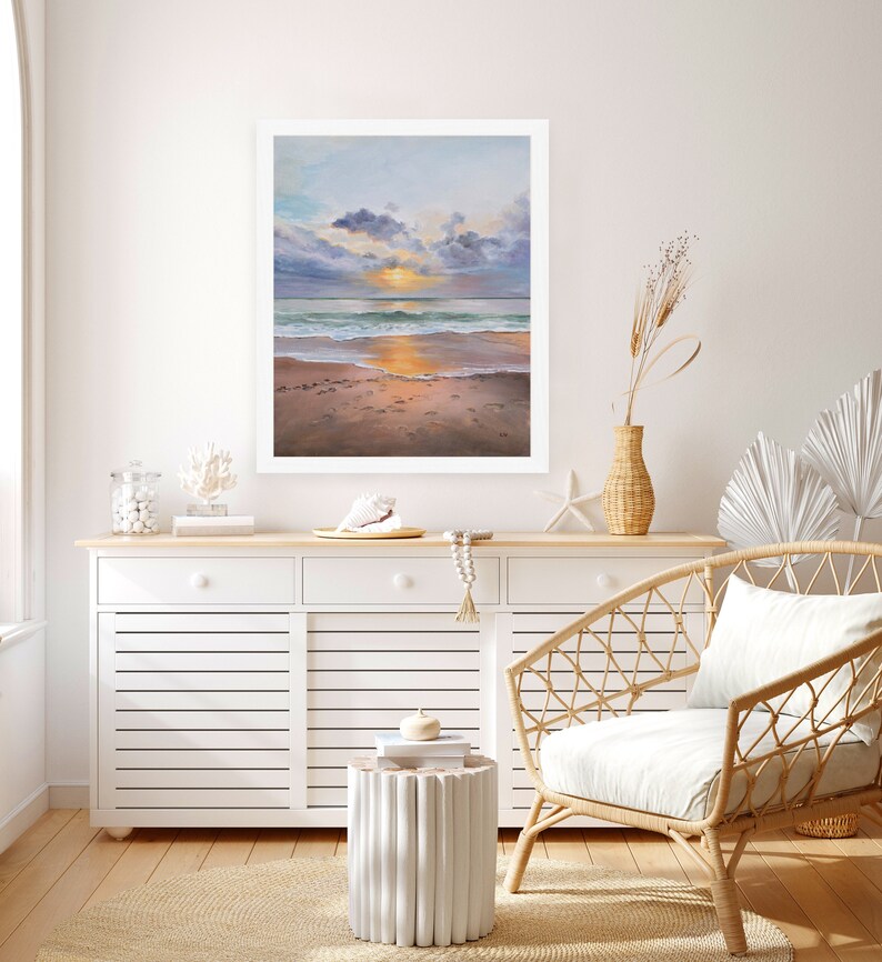 May include: A framed oil painting depicting a sunset over the ocean with soft clouds, gentle waves, and a sandy beach with footprints. The artwork is displayed on a wall above a white wooden cabinet featuring slatted doors and drawers.