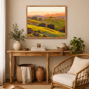 Peut inclure: Une peinture de paysage rectangulaire dans un cadre en bois clair repr&eacute;sentant des collines verdoyantes, des arbres vert fonc&eacute; et des montagnes lointaines aux teintes orang&eacute;es sous un ciel doux. L'&oelig;uvre est expos&eacute;e au-dessus d'une console.