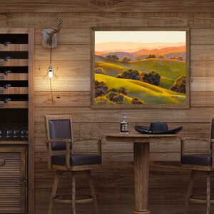 Peut inclure: Une peinture de paysage encadr&eacute;e repr&eacute;sentant des collines verdoyantes et des montagnes lointaines sous un ciel de coucher de soleil chaud, sur un mur lambriss&eacute; au-dessus d'une table ronde en bois avec un chapeau noir et une petite bouteille.