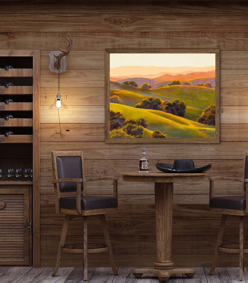 Peut inclure: Une peinture de paysage encadr&eacute;e repr&eacute;sentant des collines verdoyantes et des montagnes lointaines sous un ciel de coucher de soleil chaud, sur un mur lambriss&eacute; au-dessus d'une table ronde en bois avec un chapeau noir et une petite bouteille.