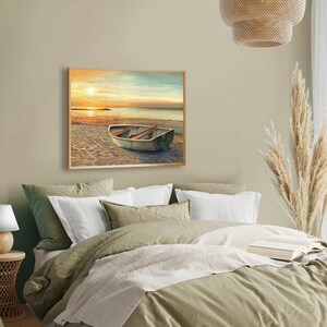 Op de afbeelding: Een rechthoekige ingelijste wandkunstprint met een landschapsschilderij van een kleine roeiboot die bij zonsondergang op een zandstrand ligt. De sc&egrave;ne toont een kalme oceaanhorizon met een felle zon, een zachte oranje lucht en een houten boot.