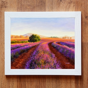 May include: A painting of a lavender field with a winding dirt road leading through the field. The field is in bloom with purple and pink flowers. The sky is a light blue with a few clouds. The painting is framed in a white frame.