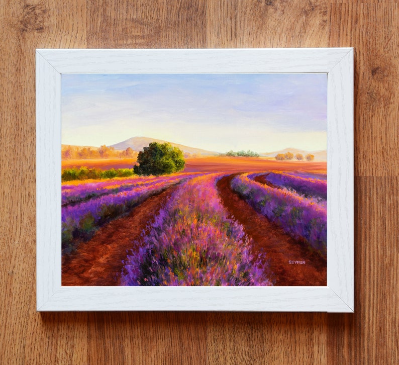 May include: A painting of a lavender field with a winding dirt road leading through the field. The field is in bloom with purple and pink flowers. The sky is a light blue with a few clouds. The painting is framed in a white frame.