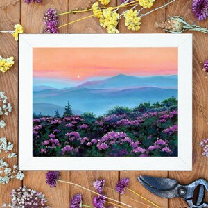 May include: A framed landscape painting depicting a sunset over blue mountain ranges with a foreground of vibrant purple wildflowers. The sky transitions from soft orange to pink hues. The artist signature SZYMER is visible in the bottom left corner.