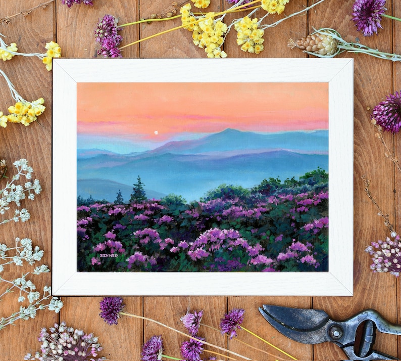 May include: A framed landscape painting depicting a sunset over blue mountain ranges with a foreground of vibrant purple wildflowers. The sky transitions from soft orange to pink hues. The artist signature SZYMER is visible in the bottom left corner.
