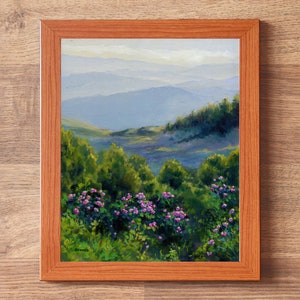 May include: A framed oil painting depicting a landscape with vibrant green bushes and clusters of small pink flowers in the foreground, leading to rolling blue-toned mountains under a soft sky. The artwork is enclosed in a light brown wooden frame.