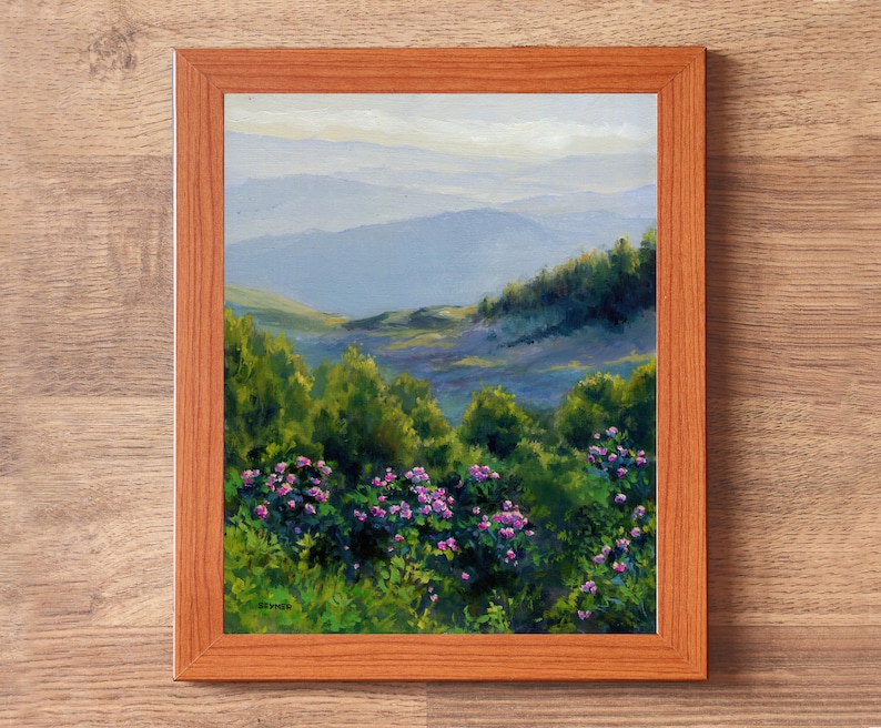 May include: A framed oil painting depicting a landscape with vibrant green bushes and clusters of small pink flowers in the foreground, leading to rolling blue-toned mountains under a soft sky. The artwork is enclosed in a light brown wooden frame.