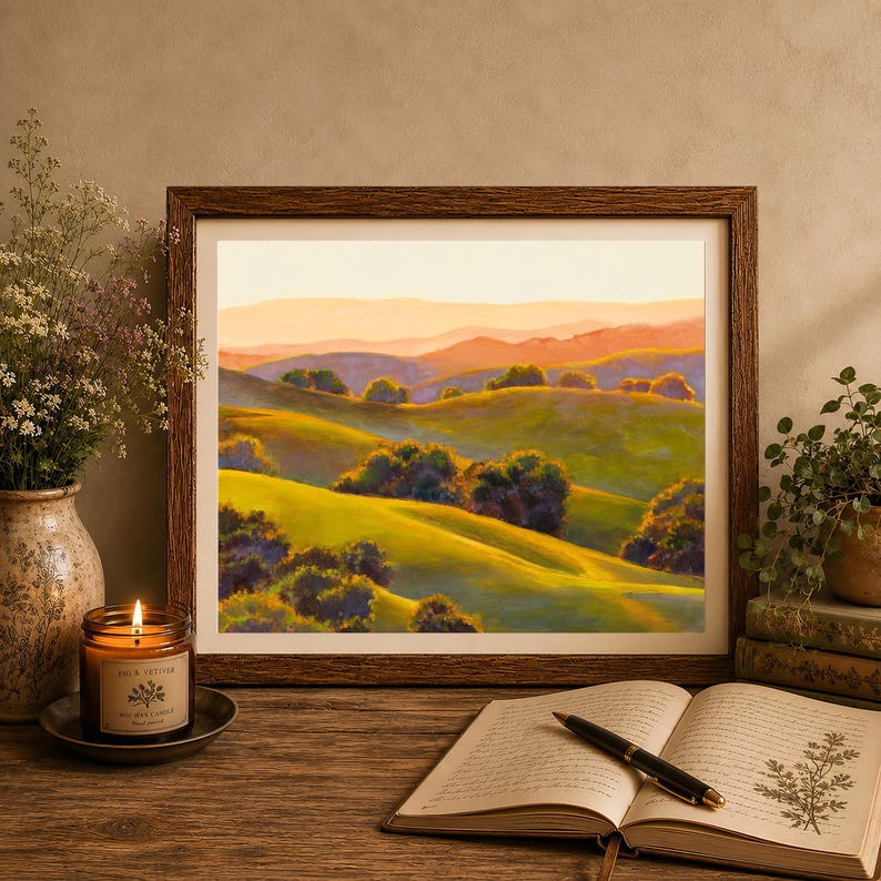 Peut inclure: Une peinture de paysage encadr&eacute;e repr&eacute;sentant des collines verdoyantes et des montagnes lointaines sous un ciel de coucher de soleil chaud. L'&oelig;uvre est pos&eacute;e sur une surface en bois avec une bougie allum&eacute;e, une plante et un livre ouvert avec un stylo.