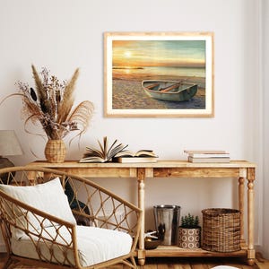 Op de afbeelding: Een ingelijste landschapsafdruk met een kleine roeiboot op een zandstrand bij zonsondergang. De sc&egrave;ne toont een kalme oceaanhorizon onder een warme, gouden lucht, met de boot op de voorgrond. Het kunstwerk is ingelijst in een licht houten frame.