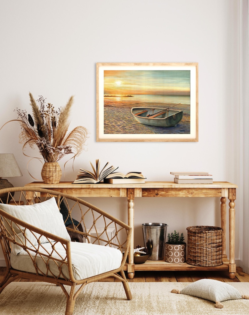 Op de afbeelding: Een ingelijste landschapsafdruk met een kleine roeiboot op een zandstrand bij zonsondergang. De sc&egrave;ne toont een kalme oceaanhorizon onder een warme, gouden lucht, met de boot op de voorgrond. Het kunstwerk is ingelijst in een licht houten frame.
