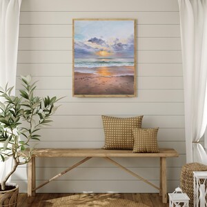 May include: A rectangular framed painting depicting a coastal sunset scene with a sandy beach, gentle ocean waves, and a sky filled with soft clouds in shades of blue, purple, and orange, with the sun glowing brightly on the horizon line.
