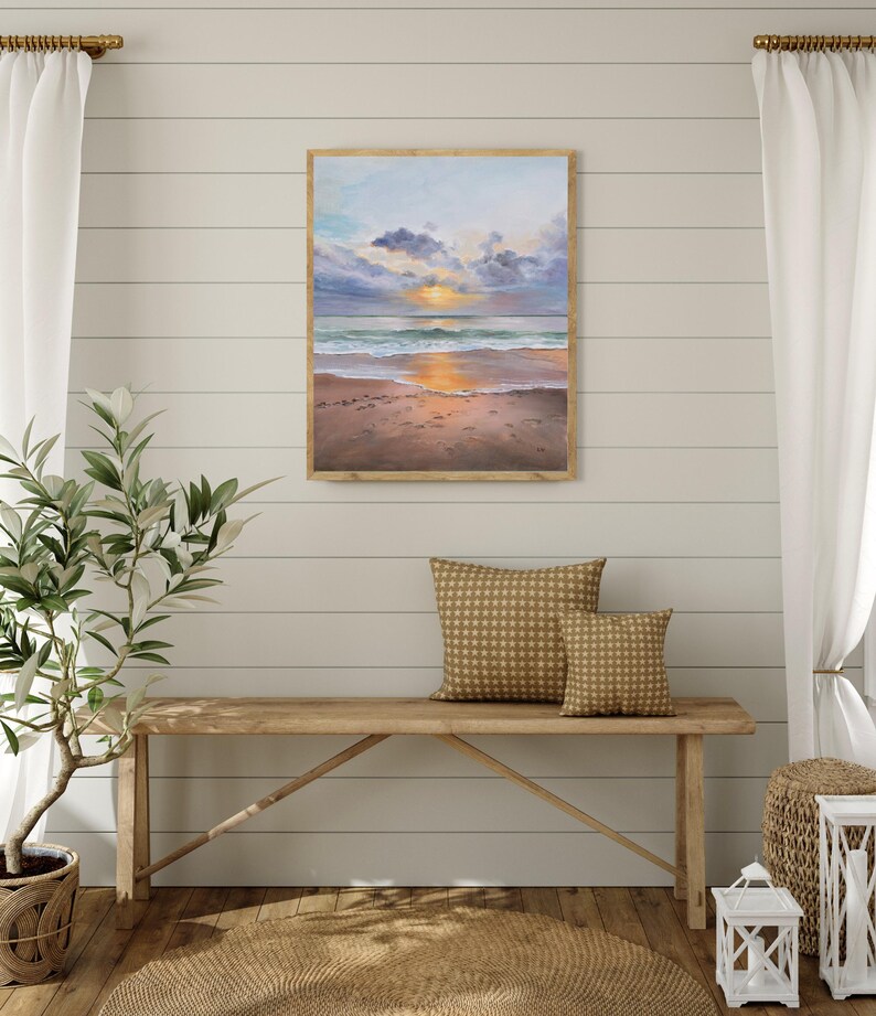 May include: A rectangular framed painting depicting a coastal sunset scene with a sandy beach, gentle ocean waves, and a sky filled with soft clouds in shades of blue, purple, and orange, with the sun glowing brightly on the horizon line.