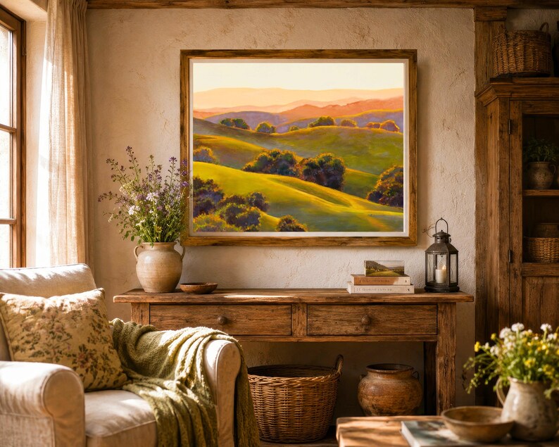 Peut inclure: Une peinture de paysage encadr&eacute;e repr&eacute;sentant des collines verdoyantes, des arbres &eacute;pars et des montagnes lointaines sous un ciel aux teintes dor&eacute;es. L'&oelig;uvre est expos&eacute;e sur un mur textur&eacute; au-dessus d'une console en bois.