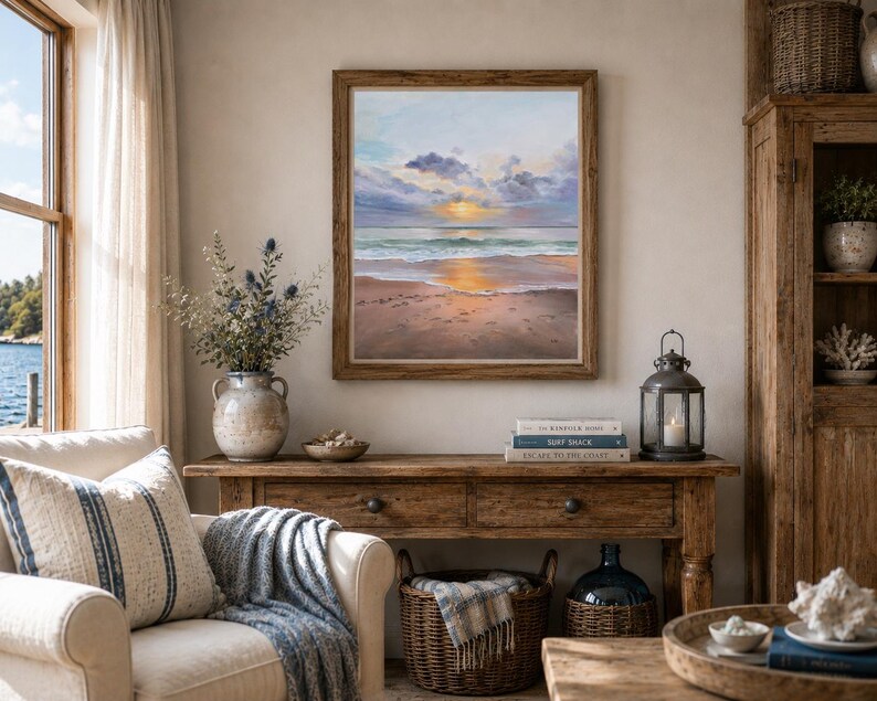 May include: A square framed oil painting depicting a sunset over a calm ocean beach with soft clouds, golden light reflecting on the wet sand and water, displayed on a wall above a wooden console table in a room with a chair and decor.