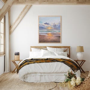 May include: A rectangular framed painting depicting a coastal sunset scene with soft clouds, a golden sun reflecting on the ocean waves, and a sandy beach in the foreground. The artwork is displayed in a light-colored wooden frame on a white wall.