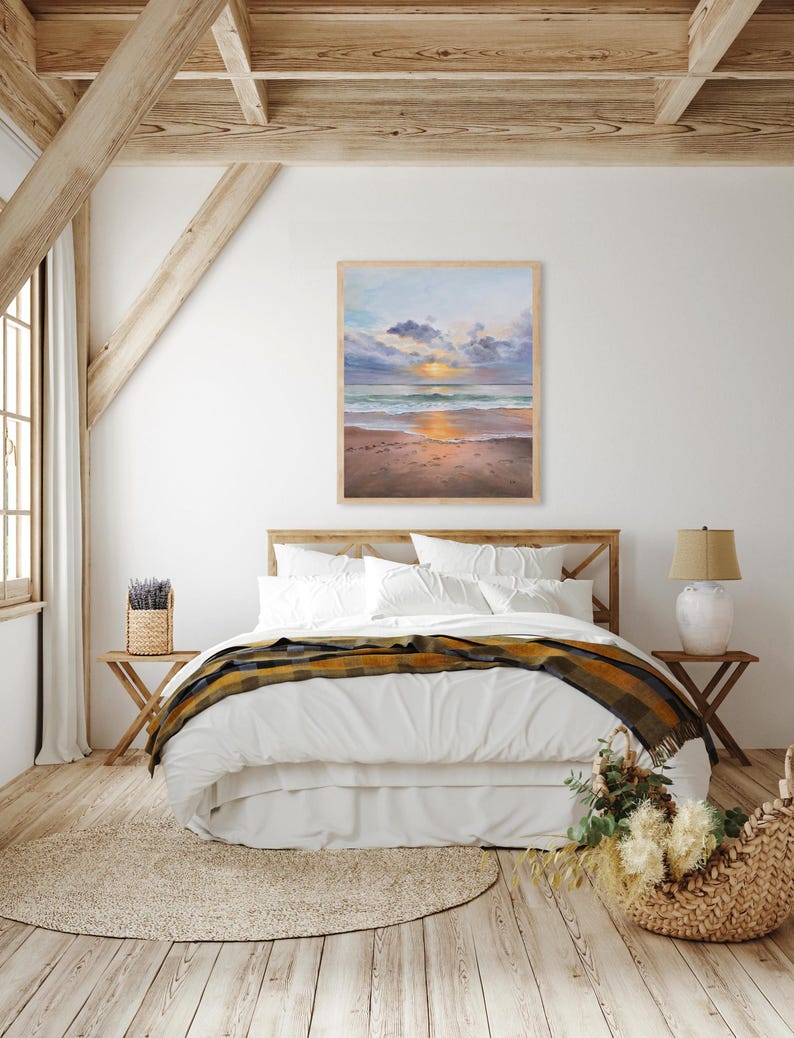 May include: A rectangular framed painting depicting a coastal sunset scene with soft clouds, a golden sun reflecting on the ocean waves, and a sandy beach in the foreground. The artwork is displayed in a light-colored wooden frame on a white wall.