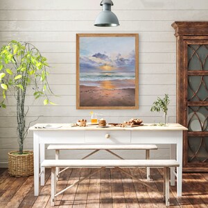May include: A framed oil painting of a sunset over a beach with soft clouds and golden light reflecting on the wet sand. The artwork is displayed on a white shiplap wall above a wooden table with benches, a potted plant, and a tall cabinet.