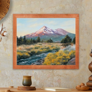 May include: A framed landscape painting shows a mountain with a snow-covered peak, a river, and yellow wildflowers. The artwork is in a light brown wooden frame. The painting evokes nature and tranquility. The frame is a warm, natural tone, adding to the rustic feel.