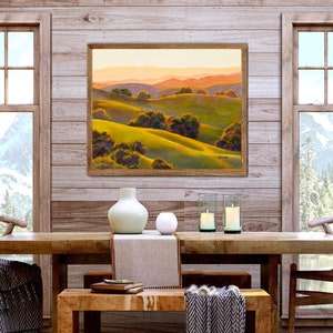 Peut inclure: Une peinture de paysage encadr&eacute;e repr&eacute;sentant des collines verdoyantes, des arbres vert fonc&eacute; et des montagnes lointaines aux teintes orang&eacute;es sous un ciel doux. L'&oelig;uvre est expos&eacute;e sur un mur en bois au-dessus d'une table &agrave; manger en bois.