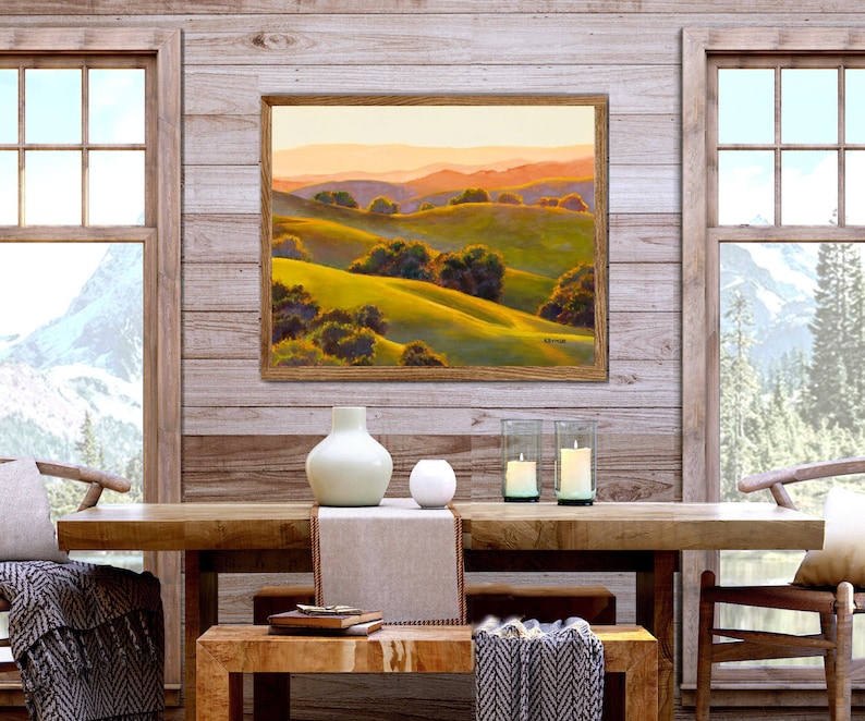 Peut inclure: Une peinture de paysage encadr&eacute;e repr&eacute;sentant des collines verdoyantes, des arbres vert fonc&eacute; et des montagnes lointaines aux teintes orang&eacute;es sous un ciel doux. L'&oelig;uvre est expos&eacute;e sur un mur en bois au-dessus d'une table &agrave; manger en bois.