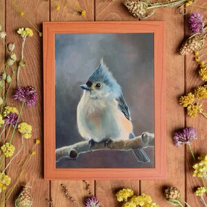 May include: A framed painting of a blue and white tufted titmouse perched on a branch. The bird is facing the left side of the frame and has a light blue head and a white body with a light brown tail. The background is a dark gray.