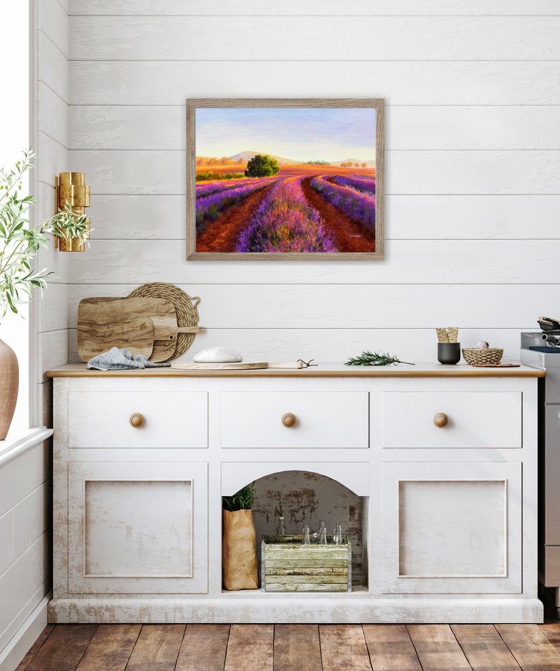 May include: A painting of a lavender field with a winding path leading to a small tree in the distance. The field is a vibrant purple and the sky is a soft blue. The painting is framed in a light wood frame.