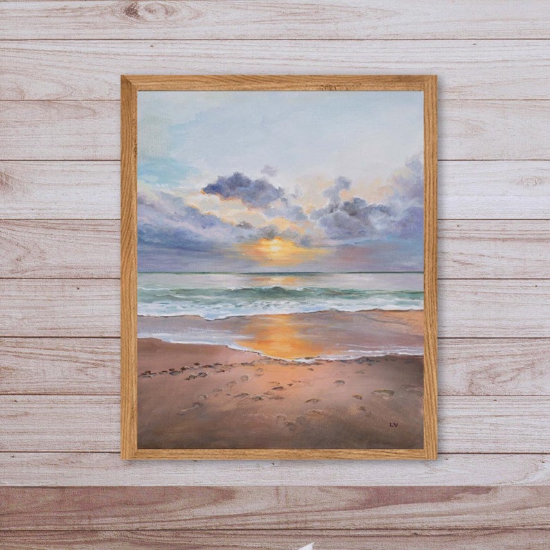 May include: An oil painting in a light wood frame depicting a sunset over the ocean. The scene features dark purple and grey clouds above a bright yellow sun, with waves washing onto a sandy beach marked by footprints and a golden reflection.