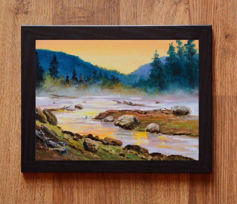 May include: An oil painting in a black frame depicting a river landscape at sunset. The scene features dark blue mountains, evergreen trees, and a misty river with rocks and driftwood. The sky is a warm orange, and the artist signature SEYMER is visible in the lower left corner.