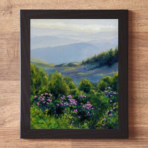 May include: A framed oil painting depicting a landscape with vibrant green bushes and clusters of small pink flowers in the foreground, leading to rolling blue mountains in the distance under a soft sky. The artwork is enclosed in a black frame.