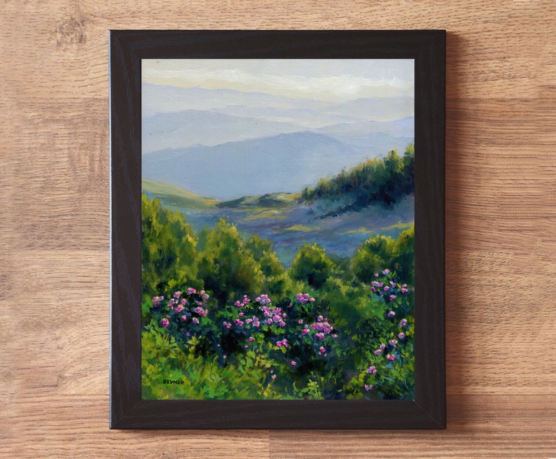 May include: A framed oil painting depicting a landscape with vibrant green bushes and clusters of small pink flowers in the foreground, leading to rolling blue mountains in the distance under a soft sky. The artwork is enclosed in a black frame.