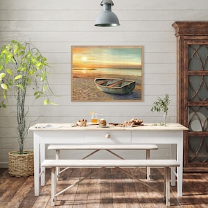 Op de afbeelding: Een rechthoekige wandkunstprint met een landschapsschilderij van een kleine roeiboot die bij zonsondergang op een zandstrand ligt. De sc&egrave;ne toont zachte oranje en gele tinten in de lucht die reflecteren op het kalme water van de zee.