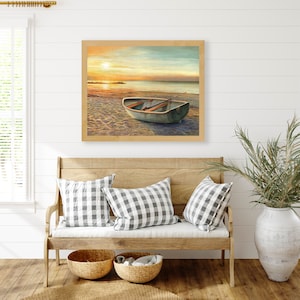 Op de afbeelding: Een ingelijst landschapsschilderij met een kleine roeiboot op een zandstrand bij zonsondergang. De sc&egrave;ne toont een kalme oceaanhorizon onder een zachte, gouden lucht met warm licht dat reflecteert op het water en het zand van het strand.