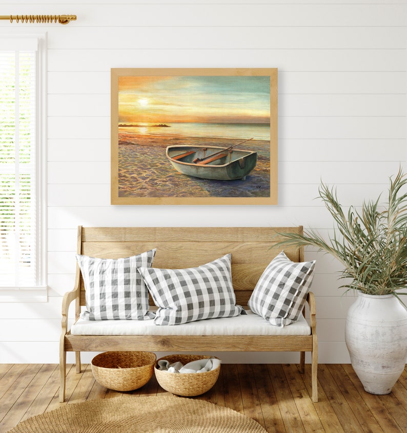 Op de afbeelding: Een ingelijst landschapsschilderij met een kleine roeiboot op een zandstrand bij zonsondergang. De sc&egrave;ne toont een kalme oceaanhorizon onder een zachte, gouden lucht met warm licht dat reflecteert op het water en het zand van het strand.