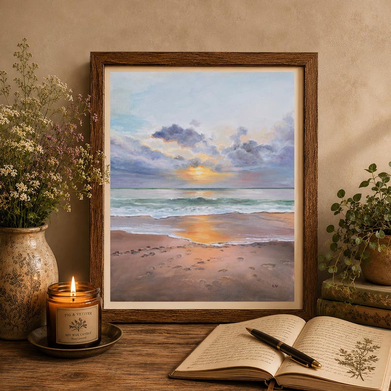 May include: A framed painting of a beach at sunset with soft clouds, calm waves, and golden light reflecting on the wet sand. The artwork is displayed in a dark wood frame, positioned next to a lit candle, a vase of flowers, and an open book.