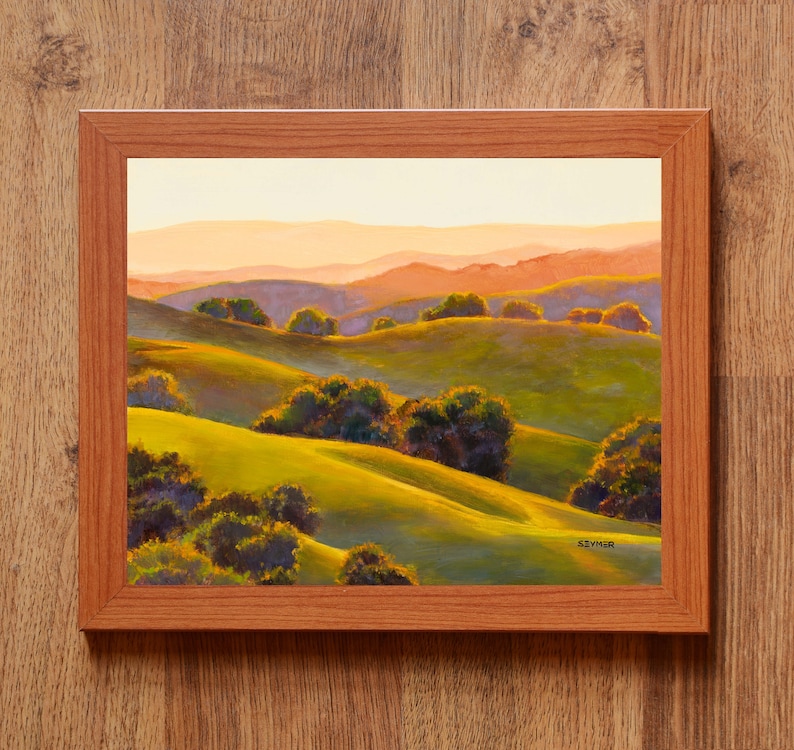 Peut inclure: Une peinture de paysage encadr&eacute;e repr&eacute;sentant des collines verdoyantes sous un ciel de coucher de soleil doux. La sc&egrave;ne pr&eacute;sente des groupes d'arbres vert fonc&eacute; dispers&eacute;s sur les pentes, avec des montagnes orange et violettes au loin. La signature SEYMER est visible.