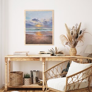 May include: A framed rectangular painting depicting a beach scene at sunset with a golden sun, blue and purple clouds, turquoise ocean waves, and wet sand. The artwork is displayed on a wall above a wooden console table with various home decor items.