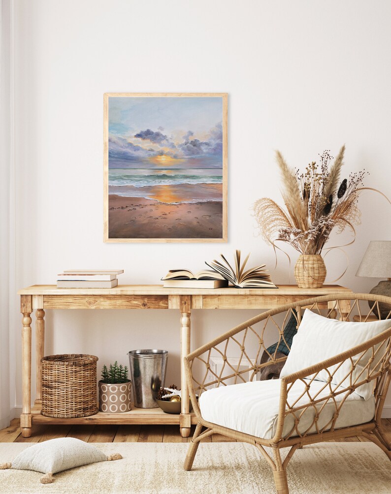 May include: A framed rectangular painting depicting a beach scene at sunset with a golden sun, blue and purple clouds, turquoise ocean waves, and wet sand. The artwork is displayed on a wall above a wooden console table with various home decor items.