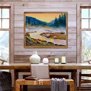May include: A framed landscape oil painting depicting a river flowing through a misty valley with dark green pine trees, rocky banks, and a golden sunset sky, displayed on a light wood paneled wall above a dining table with decor.