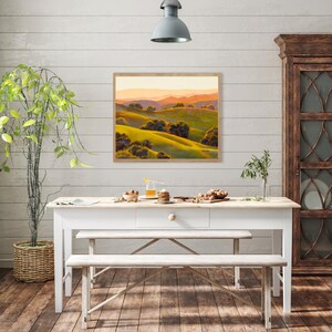 Peut inclure: Une peinture de paysage rectangulaire dans un cadre en bois clair repr&eacute;sentant des collines verdoyantes, des arbres vert fonc&eacute; et des montagnes lointaines aux teintes orang&eacute;es sous un ciel doux, accroch&eacute;e sur un mur blanc au-dessus d'une table.