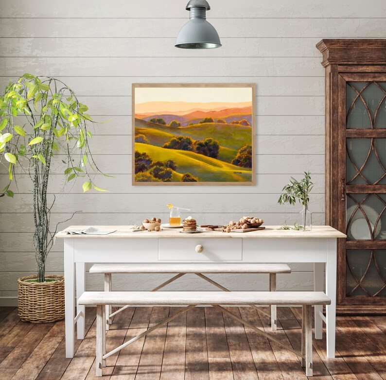 Peut inclure: Une peinture de paysage rectangulaire dans un cadre en bois clair repr&eacute;sentant des collines verdoyantes, des arbres vert fonc&eacute; et des montagnes lointaines aux teintes orang&eacute;es sous un ciel doux, accroch&eacute;e sur un mur blanc au-dessus d'une table.