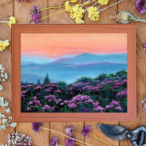 May include: A framed landscape painting depicting a sunset over blue mountain ranges with a foreground of purple wildflowers. The sky transitions from orange to pink hues. The artist signature SIYMER is visible in the lower left corner of the painting.