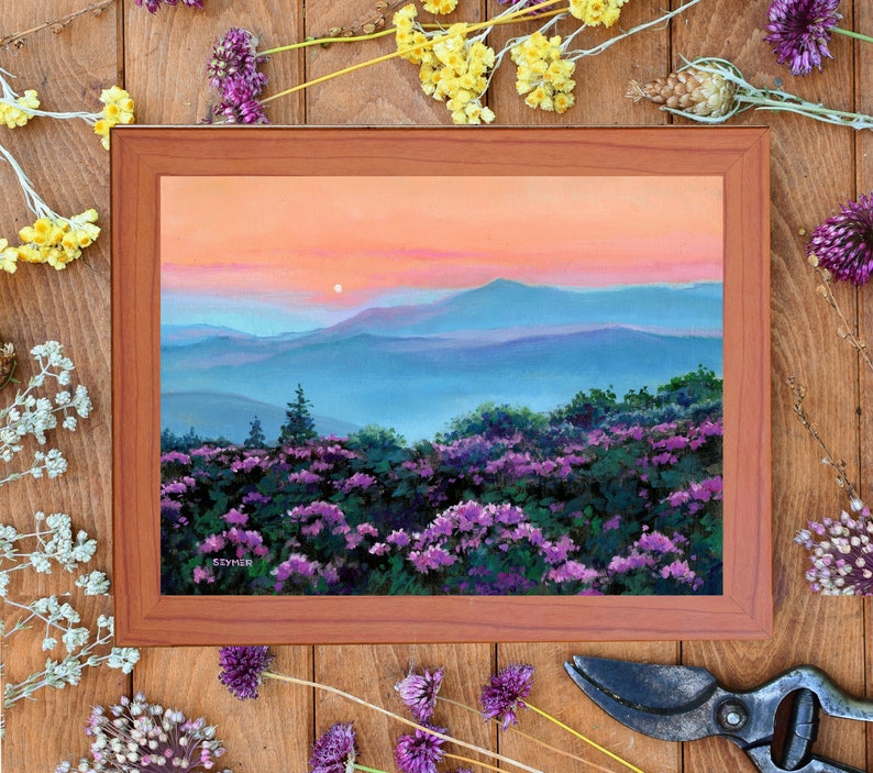 May include: A framed landscape painting depicting a sunset over blue mountain ranges with a foreground of purple wildflowers. The sky transitions from orange to pink hues. The artist signature SIYMER is visible in the lower left corner of the painting.