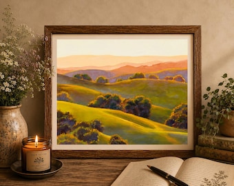May include: A framed landscape painting of rolling green hills and distant mountains under a warm sunset sky. The artwork is displayed alongside a lit candle in a glass jar, a bouquet of dried flowers in a vase, and an open book with a black pen.